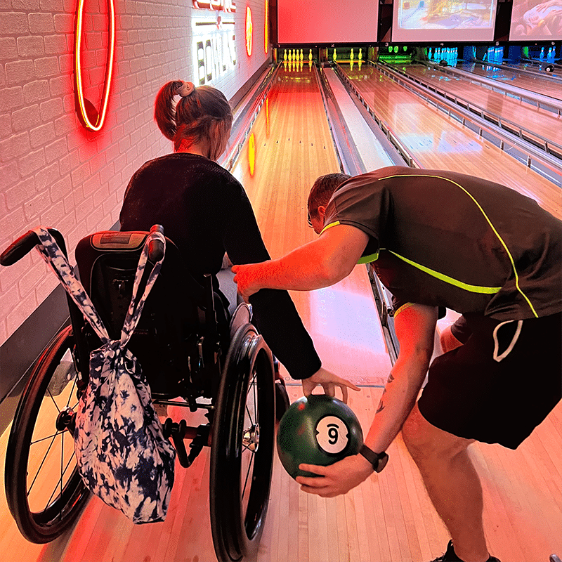 A girl in a wheelchair holding a bowling ball about to bowl with the help of a support worker